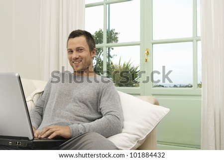 Portrait of a young professional man using a laptop while sitting on a sofa at home, smiling at camera.