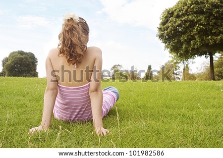 Back view of a young girl sitting down on green grass in the park, looking at the horizon.