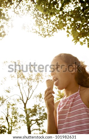 Portrait of a young girl eating an ice cream in the park, under golden light and trees.