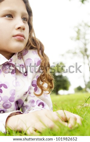 Portrait of a young girl laying down on green grass in the park, looking ahead.