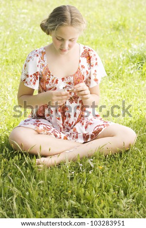 Girl pulling petals off a daisy flower while sitting on a green field, playing love me, love me not.