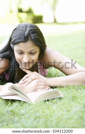 Indian girl laying down on green grass in the park, reading a book and smiling.