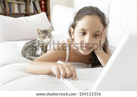 Young girl using a laptop computer at home while laying down on a white sofa with her cat.