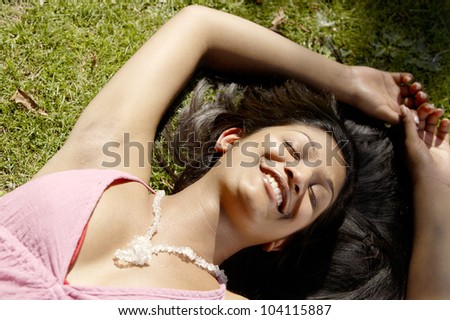 Over head portrait of a young indian woman laying down on green grass in the park, smiling.
