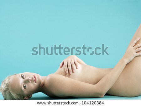 Young blond woman laying down naked on a blue background, covering herself and looking at camera.