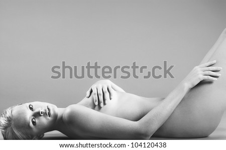 Black and white image of a naked woman laying down on a grey background, covering herself and looking at camera.