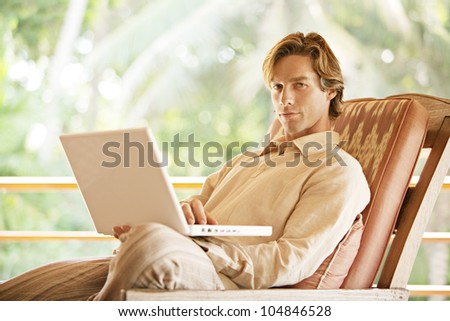 Attractive young man using a laptop computer while sitting on a tropical garden\'s balcony.