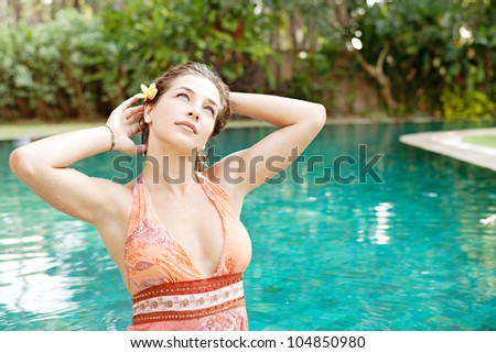 Portrait of a young woman in a swimming pool in a tropical garden, wearing a pink dress and placing a flower in her hair.