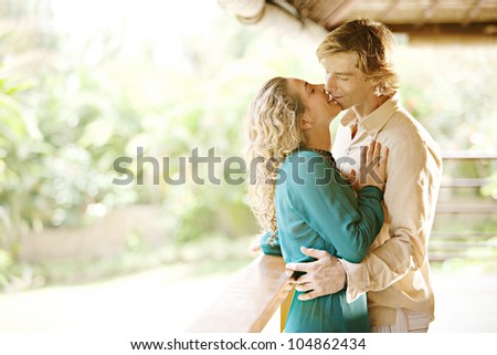 Young couple kissing on a tropical home\'s balcony overlooking a lush garden while on honeymoon.