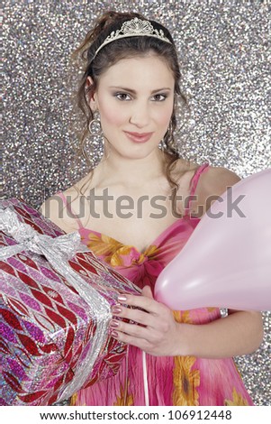 Young woman holding a gift and a balloon, and smiling on a silver glitter background.