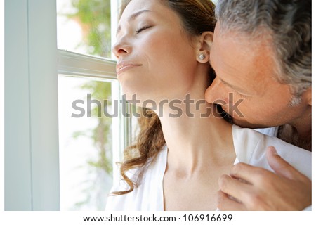 Mature man kissing woman\'s neck by large french window.