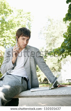 Attractive young businessman using different technology while sitting on a bench in the park.