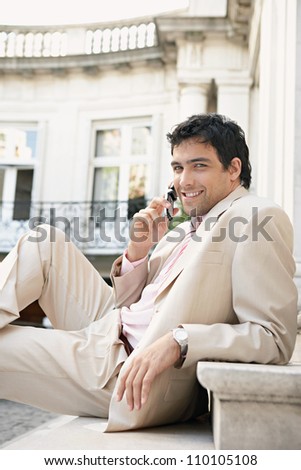Young attractive businessman smiling and using a cell phone while sitting down on stone steps in a classic building, outdoors.