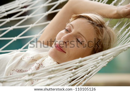 Young beautiful woman laying down on a hammock while on vacations in a tropical destination hotel, outdoors by the swimming pool and listening to music with her earphones.