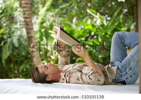 Attractive young man reading a book and laying down on an outdoors bed in a tropical lush garden while on holiday.