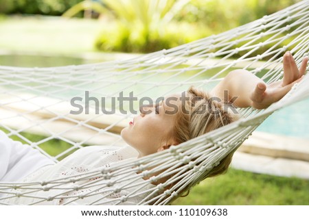 Attractive young woman laying down and relaxing on a white hammock while on vacation in a tropical garden.
