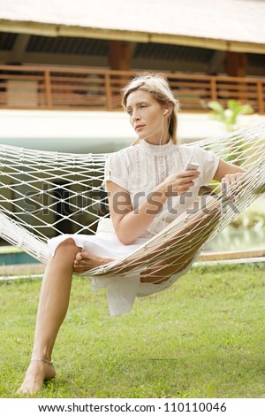 Attractive young woman listening to music while sitting on a hammock on vacation, in a tropical destination hotel\'s garden.
