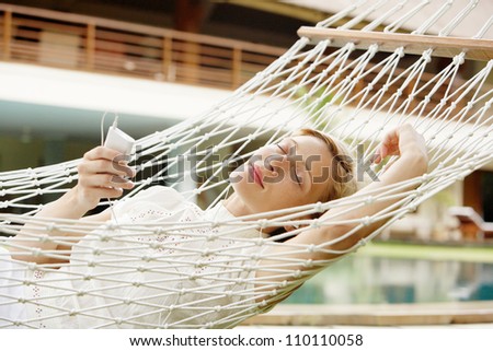 Attractive young woman laying down and relaxing on a white hammock while on vacation in a tropical garden, listening to music with her headphones and mp4 player.
