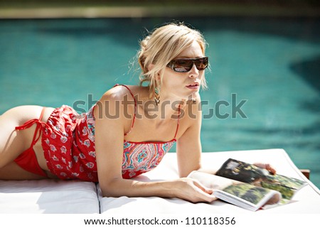 Sophisticated woman lounging by a swimming pool and reading a magazine.
