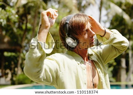 Young man listening to music with headphones while standing next to a swimming pool in a tropical garden.