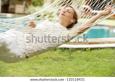 Young woman laying and relaxing on a white hammock in a tropical garden near a swimming pool, listening to music with headphones.