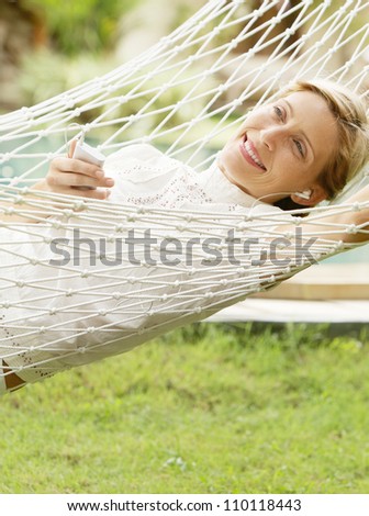 Young woman laying and relaxing on a white hammock in a tropical garden near a swimming pool, listening to music with headphones.