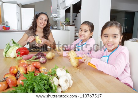 Mother and twin daughters learning to peel potatoes together in the kitchen, using a chopping board with fruit and vegetables.
