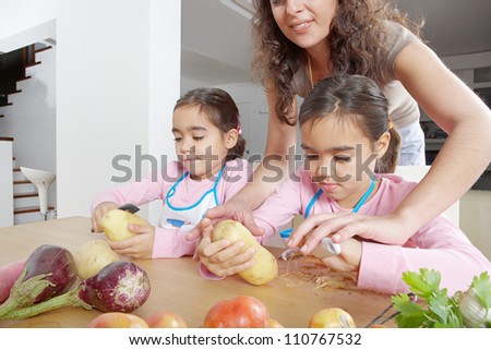 Mother teaching twin daughters to peel potatoes in the kitchen using a chopping board and peelers.