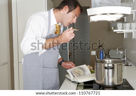 Businessman following the instructions in a cooking book to cook food at home, tasting it with a wooden spoon.