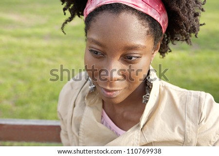 Close up portrait f a young black woman sitting on a bench with green grass in the background, smiling.