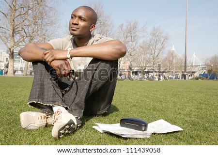 Young black tourist man sitting down on green grass in the city of London while visiting, smiling.