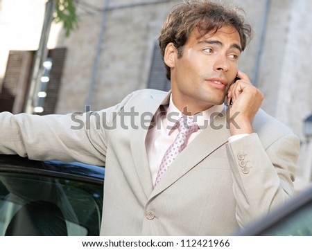 Businessman using a cell phone to make a phone call while standing by some cars in the city.