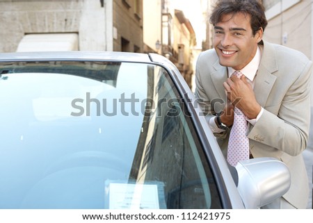Attractive young businessman grooming using a car\'s reversing mirror to tidy his tie knot, smiling.