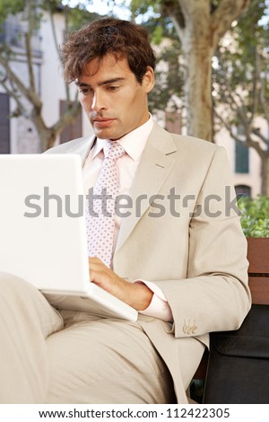 Attractive businessman sitting on a wooden bench in a classic city using a laptop computer.