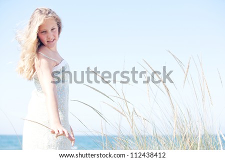 Young girl standing against a blue sky with long grass by the sea, turning and touching the grass.