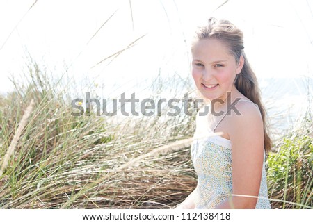 Young girl smiling and sitting down on long grass with the sea and sky in the background.