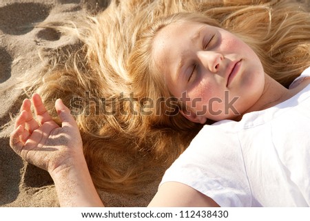 Over head view of a young girl laying down on a golden sand beach with her eyes shut while sunbathing.