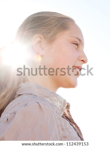 Profile close up view of a young blond girl with the sun filtering through her ponytail while she smiles against the sky.