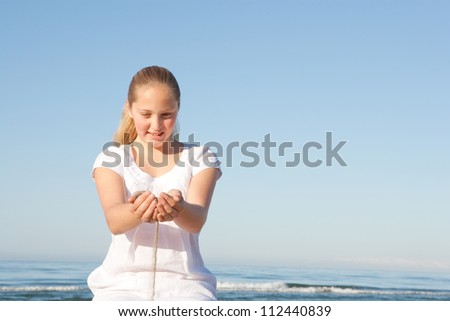 Sand filtering through a young girl\'s hands while sitting down on a beach with the sea in the background, smiling.