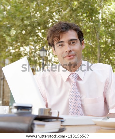 Businessman sitting at a coffee shop table using a laptop computer and smiling at camera, outdoors.
