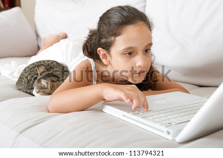 Portrait of a young girl with her sleeping cat, using a laptop computer while sitting on a white sofa at home.