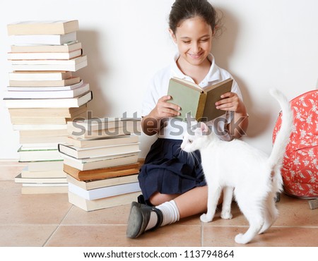 Young girl reading a book with her cat at home, sitting next to two piles of books.
