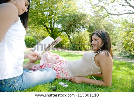 Two Indian girls spending time in the park, listening to music and reading a book, relaxing.