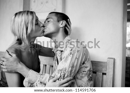 Black and white view of a young attractive couple intensely kissing while sitting down on a bench on honeymoon.