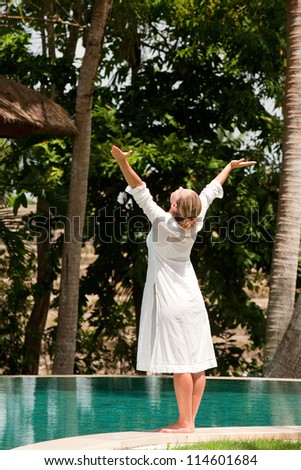 Young woman\'s figure with arms outstretched while standing at the edge of a swimming pool in a tropical nature location.