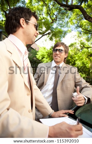 Two businessmen having a conversation while leaning on a car in a tree lined street in the city, smiling.