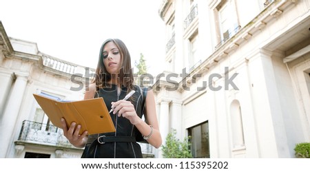Attractive businesswoman standing in a luxury classic architecture coffee shop terrace, taking notes in her agenda.