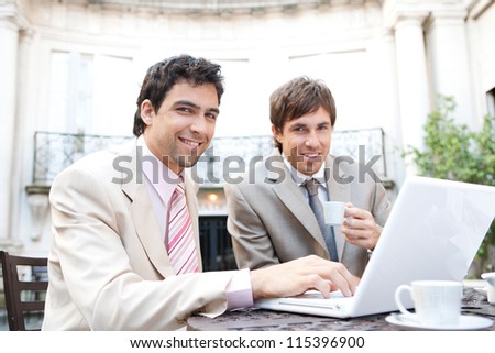 Two businessmen having a meeting while sitting in a classic coffee shop terrace, using a laptop computer and smiling at the camera.