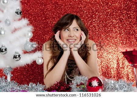 Portrait of a young attractive woman decorating a small christmas tree sitting at a table in front of a red glitter background.