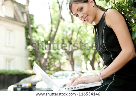 Side view of an attractive businesswoman using a laptop computer while sitting in a leafy city street during a sunny day, smiling.